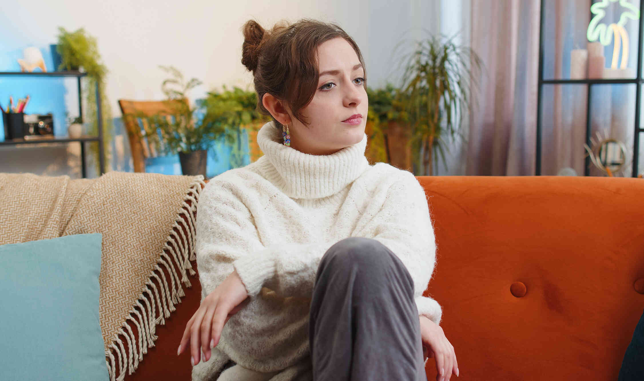 A woman in a white tutleneck sits casualy on the couch and gazes off while deep in thought.